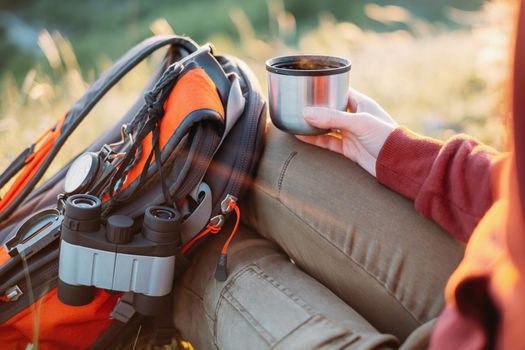 Unrecognizable explorer young woman rests with cup of tea and backpack in summer.