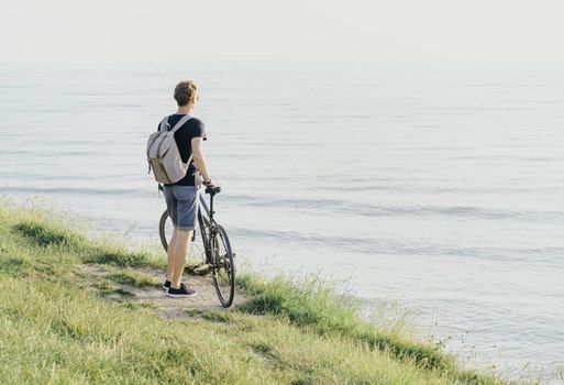Young man with backpack and bicycle standing on coast and enjoying view of sea.