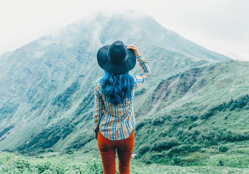 Explorer young woman in a hat standing in front of mountains in summer, rear view.