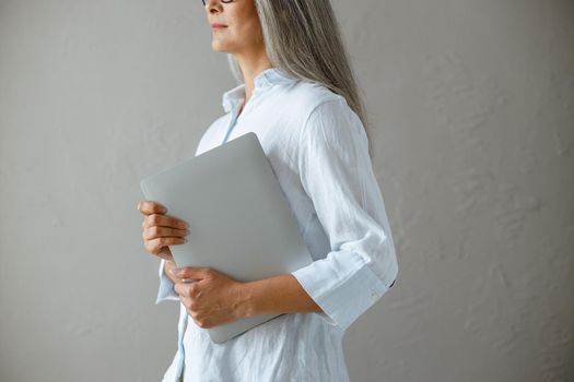 Middle aged Asian businesswoman wearing white blouse holds modern laptop standing near grey stone wall in studio side view closeup