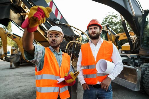 Two men engineers in workwear discussing their work standing against construction machines