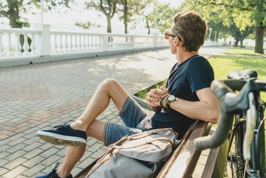 Guy rests with bicycle and backpack on bench in summer park.