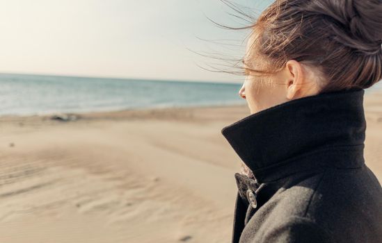 Brunette young woman wearing in coat standing on sandy coastline and looking at sea.