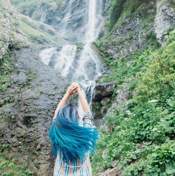 Young woman with blue hair standing in front of waterfall, her hair fluttering in the wind.