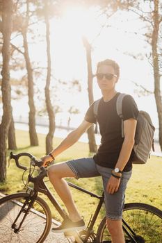 Handsome young man standing with bike in summer park at sunset.