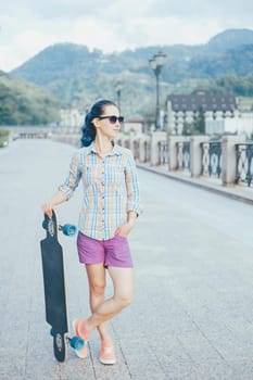 Fashionable young woman in sunglasses standing with longboard on background of mountains in summer.