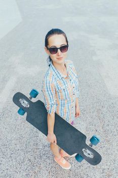 Street style girl in sunglasses standing with longboard outdoor, looking at camera.