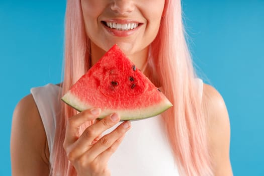 Cropped portrait of young woman with pink hair holding a slice of watermelon near her face and smiling, posing isolated over blue studio background. Beauty and health concept