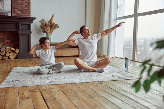 Merry little boy is training with shaply dad on mat at home and demonstrating muscles