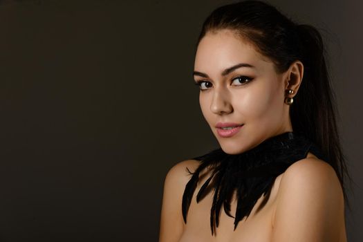 Portrait of a beautiful brunette girl with hair gathered in a ponytail with an ornament on the neck of the black feathers on dark background