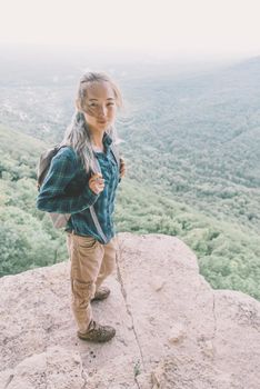 Traveler young woman with backpack standing on high cliff over summer valley, looking at camera.