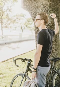 Young man standing with bicycle near a tree in summer park.