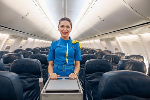 Attractive female cabin attendant in bright blue uniform smiling at camera while leading trolley cart through empty plane aisle. Travel, service, transportation, airplane concept