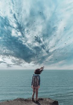 Young woman with backpack standing on coast in rainy cloudy weather, rear view.