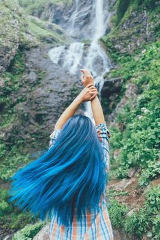 Tourist young woman with blue hair standing with raised arms in front of waterfall, concept of travel and freedom.