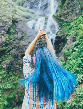 Young woman with blue hair standing with raised arms in front of waterfall in summer.