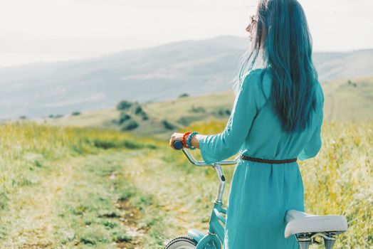 Fashionable young woman wearing in blue dress standing with bicycle on summer meadow.