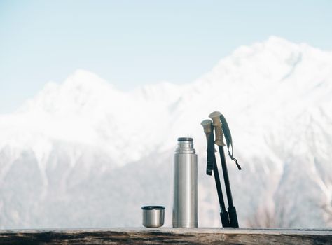 Thermos and trekking poles on wooden table on background of snowy mountains in winter.