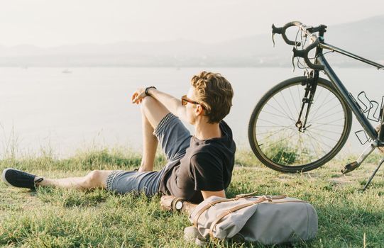 Young man with backpack and bicycle resting on summer meadow and enjoying view of sea.