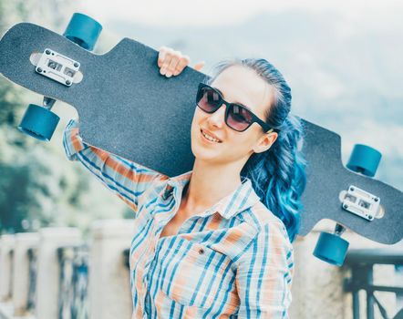 Smiling young woman in sunglasses standing with longboard outdoor, looking at camera.