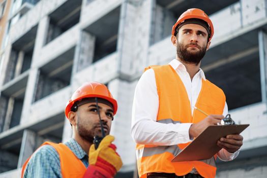 Two young male engineers in uniform and hardhats working at construction site, close up