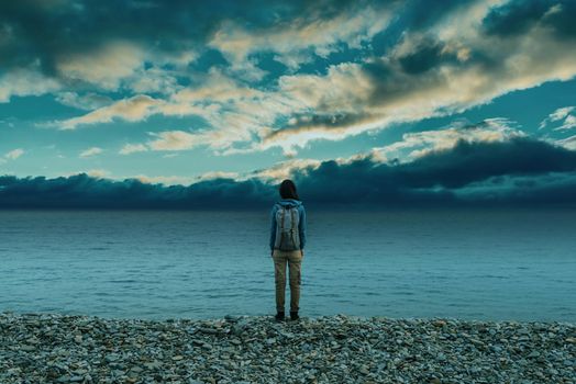 Traveler young woman with backpack standing on pebble coast near the sea and looking at cloudy sky.