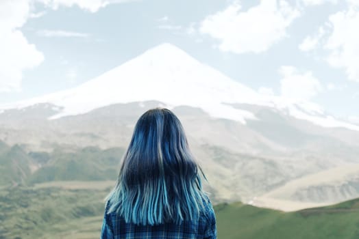 Young woman with blue hair looking at snowy mountain, rear view.