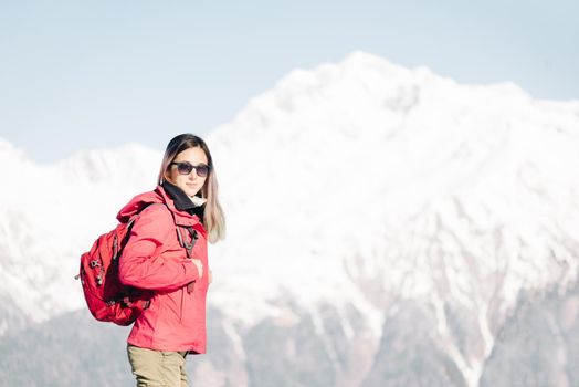 Young woman with backpack standing on background of snowy mountains, looking at camera.