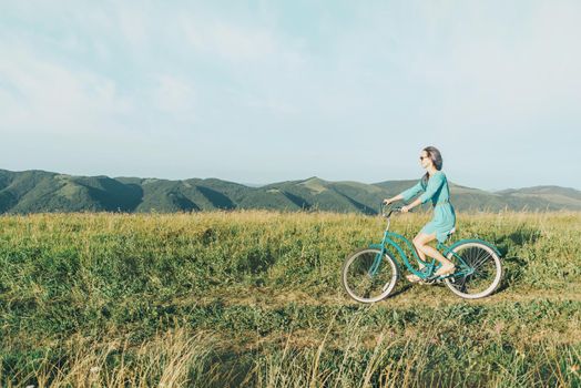 Beautiful young woman wearing in dress riding bicycle on summer meadow, side view.