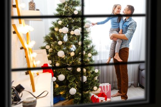 Young big family celebrating Christmas enjoying dinner, view from outside through a window into a decorated living room with tree and candle lights, happy parents eating with three kids