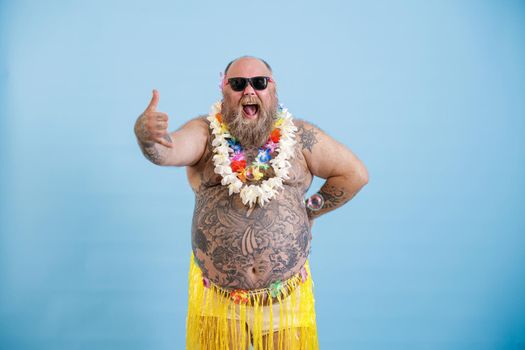 Joyful mature plump man with sunglasses wearing decorative yellow grass skirt and flowers garland shows Shaka greeting gesture on light blue background in studio