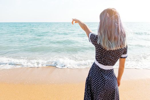 Young woman in dress standing on sand beach and pointing at sea, rear view.