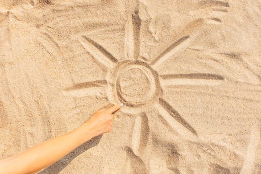 Woman draws a sun with sunbeams on the sand with her finger, view of hand.