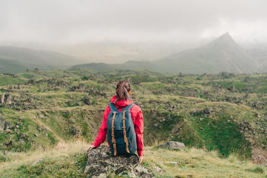 Backpacker young woman sitting on rocky stone and looking at mountains in summer, rear view.