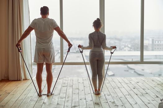 Man and woman in sportswear are standing at home and exercising with resistance bands before window