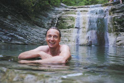 Happy young man resting in lagoon of waterfall, looking at camera.