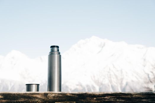 Thermos with cup on a wooden table on background of snowy mountains in winter.