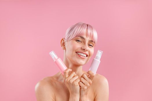 Portrait of happy young woman with pink hair holding two plastic bottles with beauty products, posing isolated over pink background. Beauty, spa, body care concept