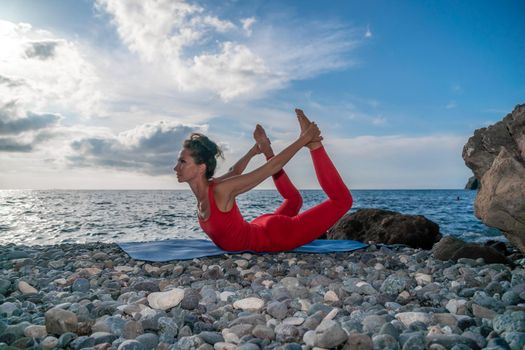 The woman in a red suit practicing yoga on stone at sunrise near the sea. Young beautiful girl in a red bathing suit sits on the seashore in lotus position. Yoga. Healthy lifestyle. Meditation