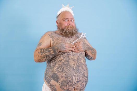 Funny man with overweight and tattoos on bare abdomen wearing toy crown covers breasts by hands holding magic stick on light blue background in studio