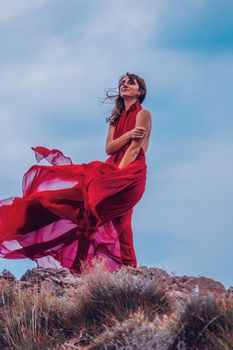 Side view a Young beautiful sensual woman in a red long dress posing on a rock high above the sea during sunrise. Girl on the nature on blue sky background. Fashion photo.