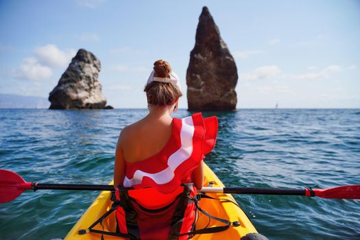 Young brunette woman in red swimsuit and Santa hat, swimming on kayak around basalt rocks like in Iceland. Back view. Christmas and travel concept