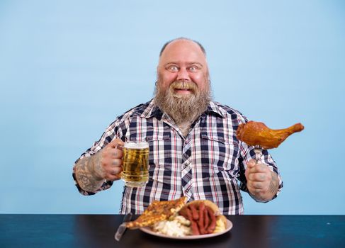 Happy middle aged plump man holds chicken leg and mug of fresh beer sitting at table with greasy food on light blue background in studio