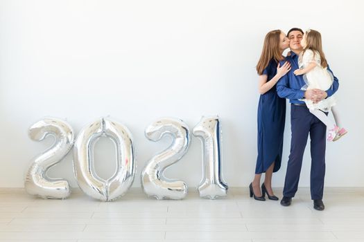 Young happy family mother and father and daughter standing near balloons shaped like numbers 2021 on white background. New year, Christmas and holiday.