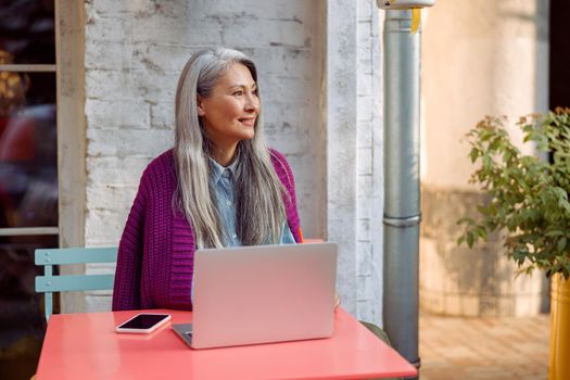 Attractive senior Asian lady in knitted jacket with smartphone and laptop sits at coral table on outdoors cafe terrace