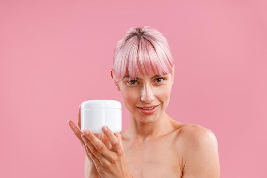 Portrait of woman with pink hair looking at camera, showing white jar with moisturizing body lotion after shower isolated over pink background. Beauty, spa, body care concept