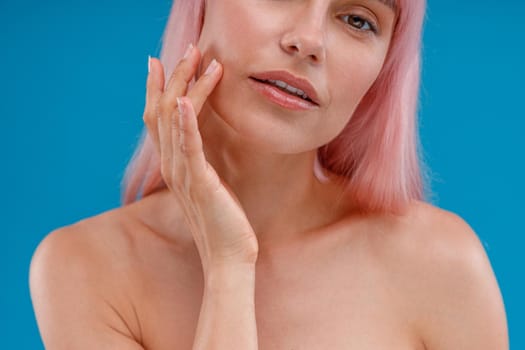 Close up shot of pure glowing skin of woman with pink hair touching her face, looking at camera, posing isolated over blue studio background. Beauty, skin care concept