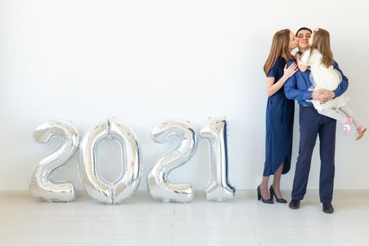 Young happy family mother and father and daughter standing near balloons shaped like numbers 2021 on white background. New year, Christmas and holiday.