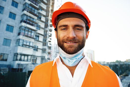 Portrait of man builder in workwear and hardhat wearing medical mask, close up photo