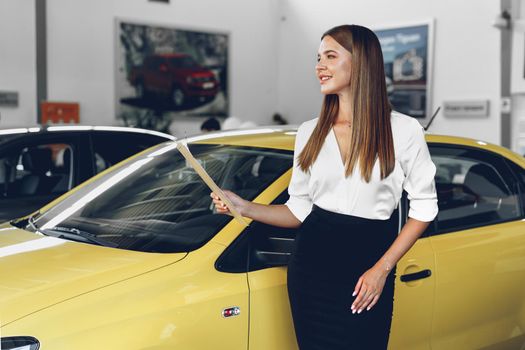 Attractive young female car dealer standing in showroom near a new car